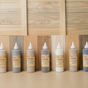 May include: A collection of Dixie Belle gel stains in various colors, displayed in squeeze bottles with white nozzles. The bottles are arranged in a row against a wooden backdrop, showcasing a range of shades from light to dark.