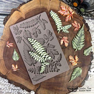 May include: A gray silicone mold with various leaf designs, alongside several painted leaf embellishments in shades of green, orange, and white. The mold features detailed botanical patterns, perfect for crafting projects. The leaves are arranged on a wooden surface.