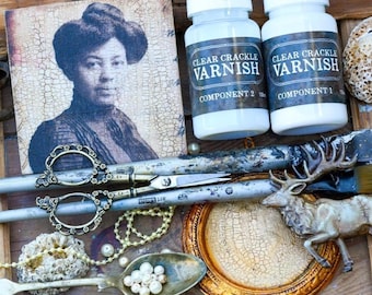 May include: Two white plastic bottles labeled Clear Crackle Varnish Component 1 and 2, a square print of a person in a hat, ornate metal scissors, a metal spoon with white beads, a gold circular frame, and a deer figurine on a wooden surface.