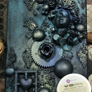 May include: A mixed media art piece on canvas with a blue and green color scheme. The piece features a variety of textures, including metal gears, beads, and a sculpted face. The piece is finished with a layer of green wax, which gives it a shimmering effect. The wax jar is labeled "Green Brocade" and "Opal Magic Wax".