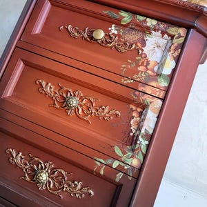 May include: A reddish-brown wooden chest of drawers featuring three horizontal drawers. Each drawer is adorned with a central gold-toned ornate metal applique and a floral botanical decal on the right side, showcasing fruits and flowers.