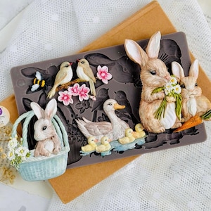 May include: A silicone mold featuring various spring-themed designs. Includes a bunny in a basket, bunnies with flowers and a carrot, birds on a branch, a duck with ducklings, and a bee. The mold is a dark gray color.