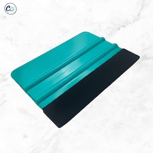 May include: A rectangular vinyl application squeegee featuring a bright teal plastic handle with two raised horizontal ridges for grip and a wide, flat black felt edge attached to one side for smoothing surfaces during craft projects.