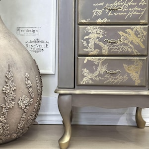 May include: A gray cabinet with gold accents. The drawers feature gold bird and floral designs, with script writing. The cabinet has curved legs with gold tips and ornate gold handles. The cabinet is next to a decorative vase.