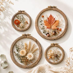 May include: Four circular embroidery hoops with faux wood grain frames containing pressed dried flowers and foliage. The arrangements feature a large orange maple leaf, wheat stalks, white and orange blossoms, and small green sprigs.