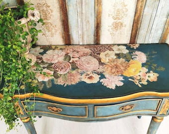 May include: A teal-colored antique console table with gold trim and floral design. The table features two drawers with ornate gold handles and curved legs. A basket of greenery is on the left side of the table.