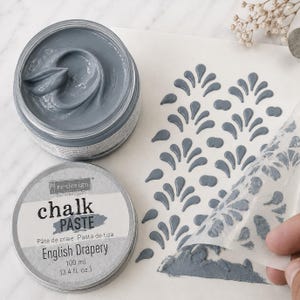 May include: A round container of blue-gray chalk paste labeled re-design with chalk paste English Drapery 100 ml 3.4 fl. oz. Next to it, a stencil is being lifted to reveal a raised, repeating teardrop pattern in the same blue-gray paste.
