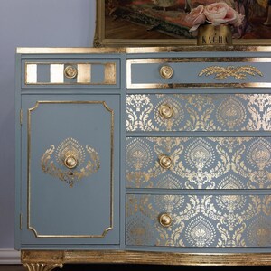 May include: A blue-gray painted sideboard with gold accents. The cabinet features gold leaf trim, drawer pulls, and decorative stenciled patterns. The top drawer has a gold stripe design. The cabinet has a classic, vintage style.