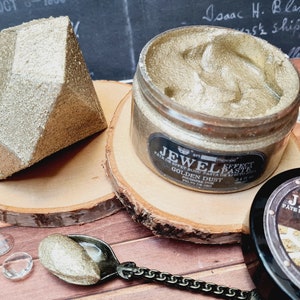 Puede incluir: Un tarro de pasta de efecto Jewel Effect en color Golden Dust, con una textura dorada met&aacute;lica. La etiqueta dice Jewel Effect Paste Golden Dust 100 ml. Al lado hay una forma geom&eacute;trica dorada facetada y una cuchara peque&ntilde;a con la pasta.