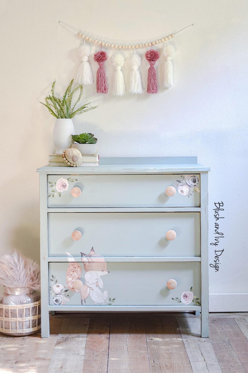May include: A light blue painted dresser with three drawers, featuring floral and woodland animal decals. The dresser has round, light pink knobs and is decorated with a garland of wooden beads and tassels. The text "Blush and Ivy Design" is on the side.