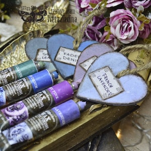 May include: A collection of small wooden heart-shaped ornaments with labels that read "French Lavender", "Faded Denim", and "Sage Leaves". The hearts are painted in shades of blue, purple, and green.  Four tubes of paint are visible in the foreground, labeled "Wax Finish" and featuring colors like "Lavender", "Pink", "Denim", and "Leaves".