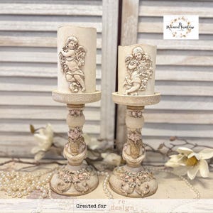 May include: Two cream-colored candle holders with cherub designs. The candle holders have a detailed, ornate base with pink floral accents. The candles themselves are cylindrical and feature a relief of a cherub. The overall style is vintage and romantic.