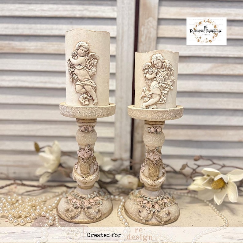 May include: Two cream-colored candle holders with cherub designs. The candle holders have a detailed, ornate base with pink floral accents. The candles themselves are cylindrical and feature a relief of a cherub. The overall style is vintage and romantic.