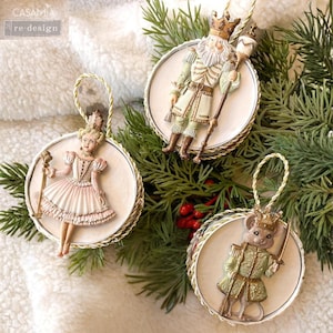 May include: Three round Christmas ornaments featuring raised figures: a nutcracker in a green uniform, a ballerina in a pink dress, and a mouse king in a green coat. Each ornament has a white circular base, gold trim, and a gold hanging cord.