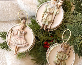 May include: Three round Christmas ornaments featuring raised figures: a nutcracker in a green uniform, a ballerina in a pink dress, and a mouse king in a green coat. Each ornament has a white circular base, gold trim, and a gold hanging cord.