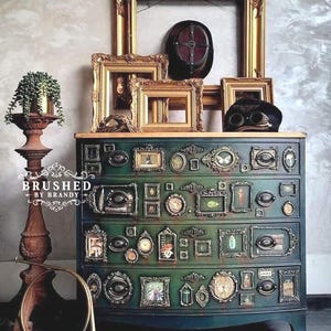 May include: A teal green dresser with multiple small framed pictures and decorative elements. The top of the dresser features several gold picture frames, a dark helmet, and goggles. A decorative stand with a plant and a brass horn are in the foreground. The text "BRUSHED BY BRANDY" is visible.