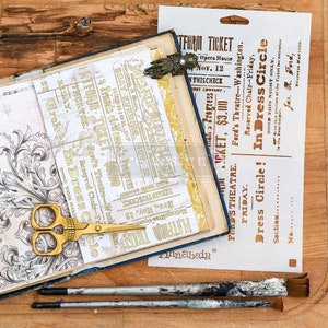 May include: An open scrapbook with vintage-style ephemera, including ticket stubs and patterned paper. A gold-colored hand-shaped clip holds the pages. Gold scissors and paintbrushes are also visible.