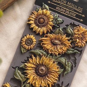 May include: A grey silicone mold for crafting, labeled Decor Moulds Golden Solstice, featuring recessed designs of multiple sunflowers with detailed petals, centers, and leaves. The mold is rectangular with a black header card attached at the top.