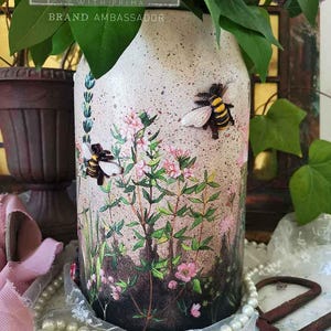 May include: A white ceramic jar with a floral design and two bees. The jar is decorated with a speckled paint finish and has the text "re.design with prima brand ambassador" on the front.