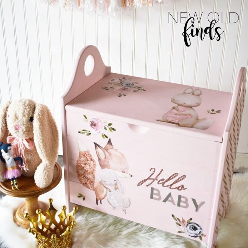 May include: A pink wooden toy chest with a handle, decorated with floral and animal illustrations. The chest features the text "Hello Baby" and is next to a stuffed bunny and a gold crown.