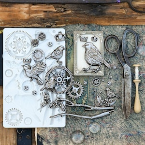 May include: A white silicone mold featuring various embossed shapes including birds, gears, crowns, and branches. Beside the mold are several metallic-colored cast pieces of birds, crowns, and gears, along with vintage metal scissors and a wooden tool.