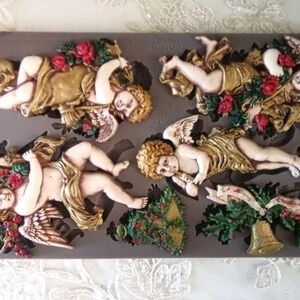 May include: A gray silicone mold featuring several cherub designs with gold-colored robes, wings, and floral accents. The cherubs are in various poses, some holding roses. A Christmas tree and bell are also included in the mold.