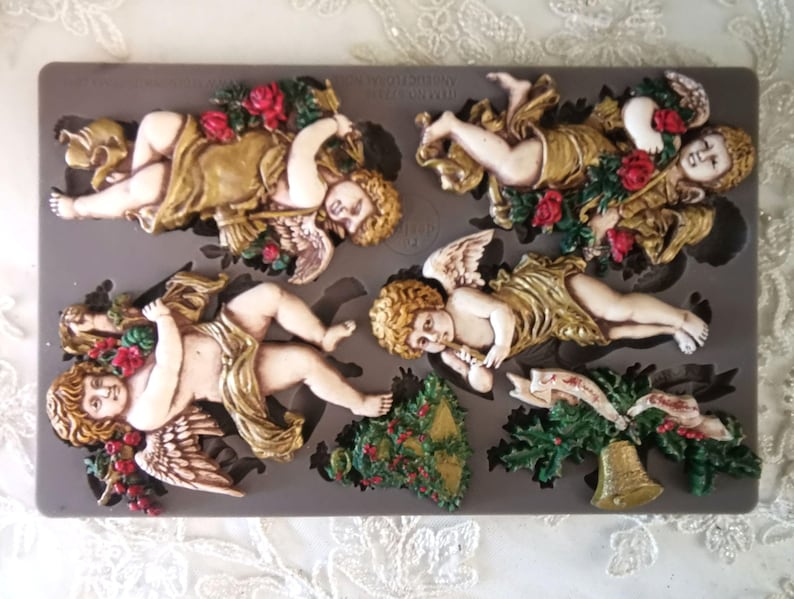 May include: A gray silicone mold featuring several cherub designs with gold-colored robes, wings, and floral accents. The cherubs are in various poses, some holding roses. A Christmas tree and bell are also included in the mold.