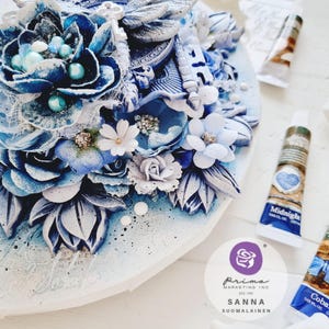 May include: A close-up of a blue and white mixed media art piece with flowers, a dragonfly, and a key. The art piece is on a white background. The text "Prima Marketing Inc. Est. 1996 Sanna Suomalainen" is visible on a white circle sticker.