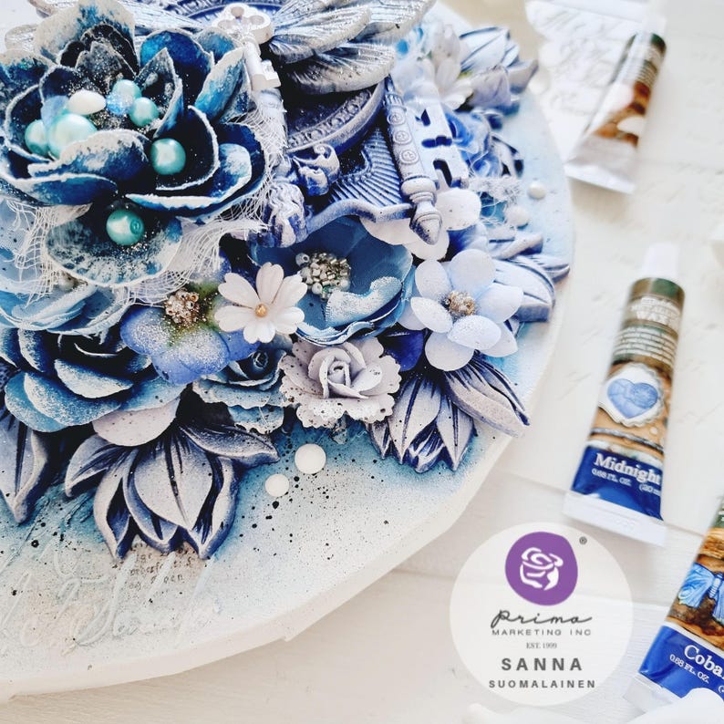 May include: A close-up of a blue and white mixed media art piece with flowers, a dragonfly, and a key. The art piece is on a white background. The text "Prima Marketing Inc. Est. 1996 Sanna Suomalainen" is visible on a white circle sticker.