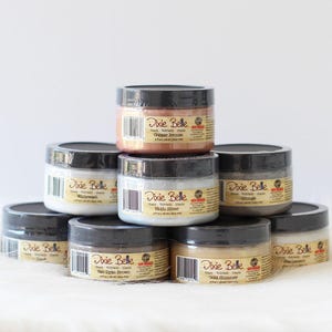 May include: A collection of Dixie Belle glaze jars in various colors, including Copper Bronze, Hello Silver, and Gold Shimmer. Each jar has a black lid and a label with the brand name and color. The jars are stacked on a white surface.