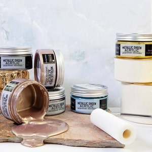 May include: A collection of metallic sheen acrylic paints in various colors, including gold, bronze, and teal, displayed in small jars with silver lids. One jar is tipped over, with the metallic paint spilling onto a wooden surface. A white paint roller is also present.