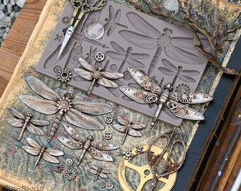 Puede incluir: Un surtido de adornos de libélulas y engranajes de estilo steampunk. La imagen incluye un molde de silicona, tijeras decorativas y libélulas acabadas con intrincados detalles mecánicos. La paleta de colores es principalmente plateada y bronce metálico.