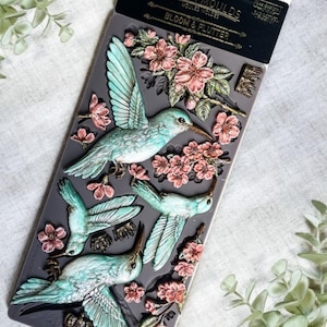 May include: A gray silicone mold featuring three hummingbirds and pink flowers. The hummingbirds are light blue with detailed wings and long beaks. The mold is labeled "BLOOM & FLUTTER" in gold lettering.