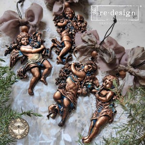 May include: Ornate bronze-colored cherub figurines with blue accents. The cherubs are in various poses, some holding flowers or playing instruments. The image includes the text "re-design with Prima Brand Ambassador".