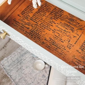 May include: Open drawer of a light blue cabinet with a gold knob. The drawer interior features a vintage-style paper design with handwritten text and the words "Thankful" and "Paris 1889".