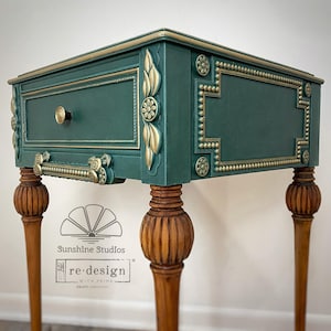 May include: A teal-colored nightstand with gold accents. The drawer features a gold knob and handle. The legs are a light brown color with carved details. The nightstand has decorative floral and geometric gold trim.