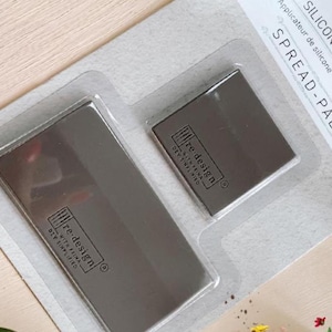 May include: A package containing two silicone applicators. The applicators are rectangular and gray, with the text "re design" printed on them. The package also includes the text "SILICONE APPLICATOR" and "SPREAD-PALS".