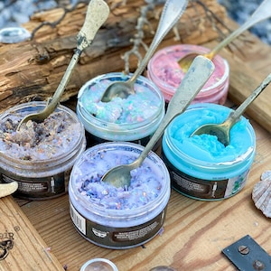 May include: Four small glass jars filled with colorful bath scrubs, each with a silver spoon resting on top. The scrubs are in shades of purple, blue, pink, and brown. The jars are on a wooden surface with a starfish and seashells.