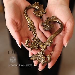 May include: A pair of ornate gold-toned metal handles in the shape of a heart. The handles have a detailed, intricate design with swirls and curves. The handles are held in the hands of a person.
