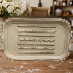 May include: An oblong, off-white decorative tray with a detailed, raised border. The tray features seven horizontal, textured designs, including rope, bead, and oval patterns. The tray is set on a wooden surface, with a blurred background of flowers and shelves.