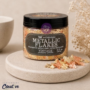 May include: A clear plastic jar with a black lid containing metallic flakes in shades of gold and copper. The label reads METALLIC FLAKES VINTAGE and 5 fl. oz. (150 ml). A small pile of the metallic flakes sits in front of the jar on a round base.