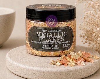 May include: A clear plastic jar with a black lid containing metallic flakes in shades of gold and copper. The label reads METALLIC FLAKES VINTAGE and 5 fl. oz. (150 ml). A small pile of the metallic flakes sits in front of the jar on a round base.