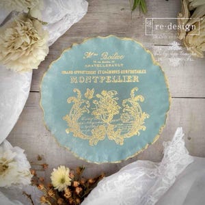 May include: A round, light blue decorative plate with a scalloped edge and gold trim. The plate features gold text and floral designs, including the word "MONTPELLIER". The plate is surrounded by white fabric and flowers.