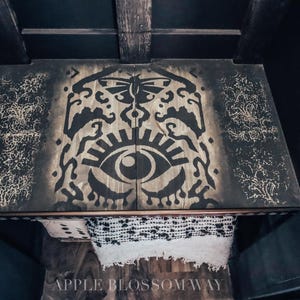 May include: A rectangular wooden table top featuring a dark, stenciled design of a central eye and a moth above it, flanked by intricate floral patterns. The wood grain is visible beneath the black paint. The text APPLE BLOSSOM WAY is visible.