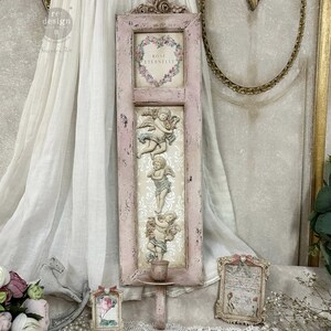 May include: A distressed pink wall decor piece featuring a framed section with the text "ROSE ETERNELLE" inside a floral heart. Below are three cherub sculptures. A small candle holder is attached at the bottom.