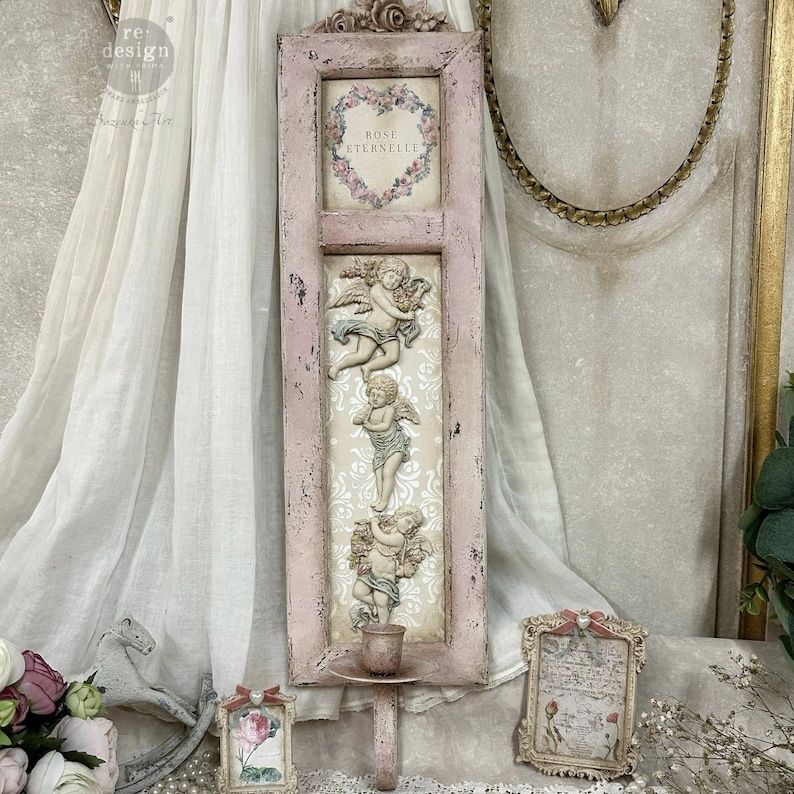May include: A distressed pink wall decor piece featuring a framed section with the text "ROSE ETERNELLE" inside a floral heart. Below are three cherub sculptures. A small candle holder is attached at the bottom.