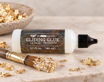 Puede incluir: Un frasco de plástico blanco de pegamento Art Extravagance Gliding Glue con tapa negra, etiquetado con 80 ml. Un pincel de madera con copos de pan de oro en las cerdas descansa junto al frasco, rodeado de trozos de pan de oro esparcidos.