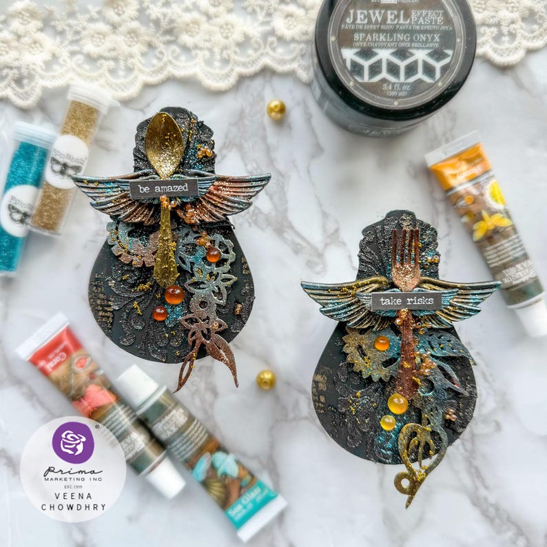 May include: Two black and gold decorative ornaments with wings, gears, and glitter. One ornament has a spoon with the text "be amazed" and the other has a fork with the text "take risks".