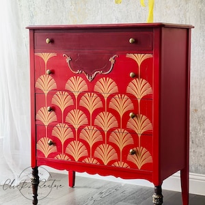 May include: A red painted dresser with gold fan-shaped designs on the drawers. The dresser has a dark red top and drawer, with gold knobs and decorative hardware. The legs are dark brown with gold accents. The dresser is a piece of furniture.