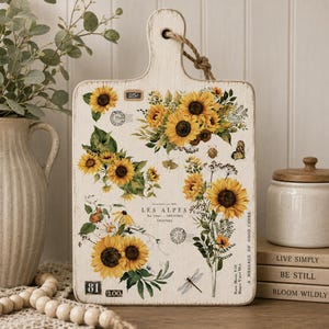 May include: A rectangular wooden cutting board with a distressed white finish and a jute rope handle. The surface features sunflower illustrations, vintage postage stamps, the text LES ALPES, and the phrase A MESSAGE OF GOOD CHEER.